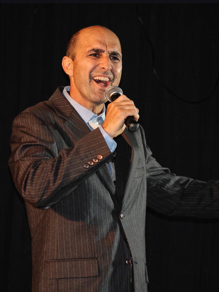 Rick in a pinstripe suit signing into a handheld microphone on a darkened stage